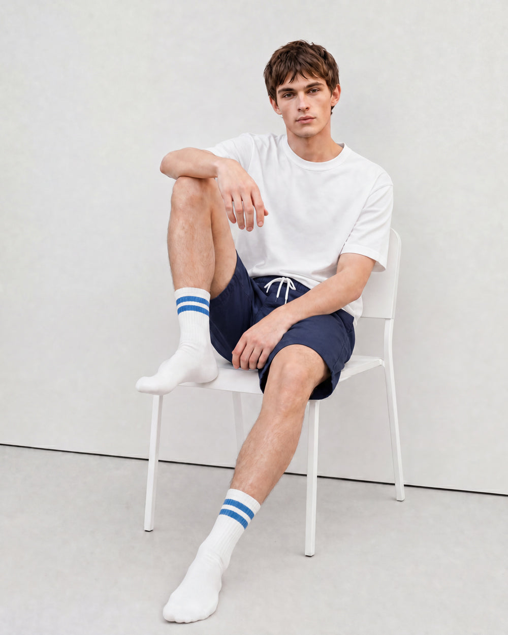 Man wearing white crew socks with blue stripes, navy shorts and a white t-shirt on a white chair