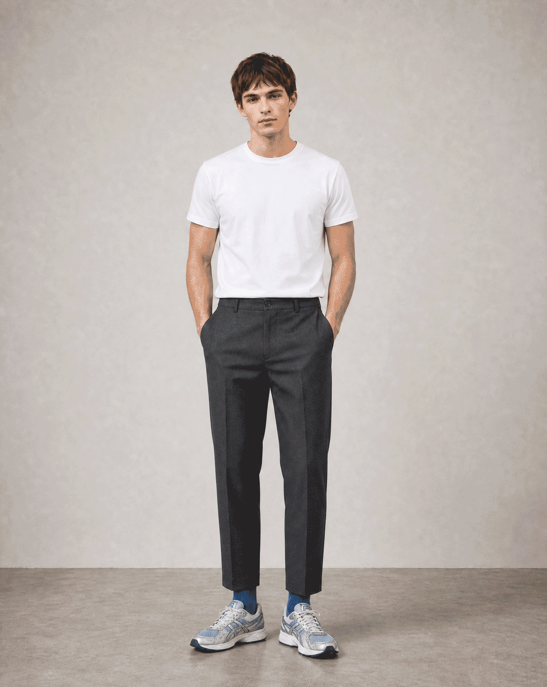 Man wearing charcoal tailored trousers, white crew-neck t-shirt and silver ASICS sneakers, with Democratique Fine Rib Socks in blue visible at the ankle.