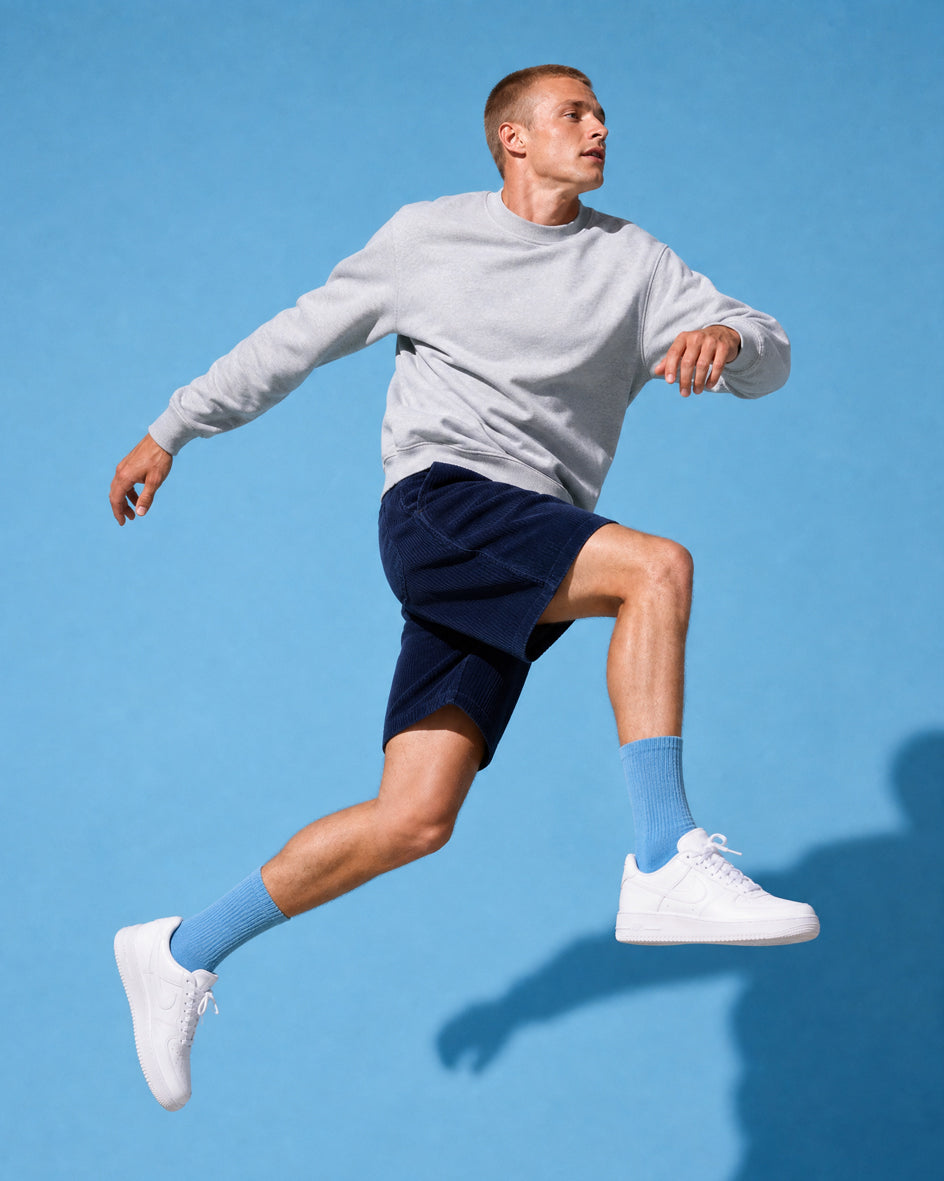 Man leaping in motion wearing navy corduroy shorts, grey marl crewneck sweatshirt, white Nike Air Force 1 sneakers and sky-blue crew socks against a bright blue background. Casual summer menswear with matching sock and background tones.