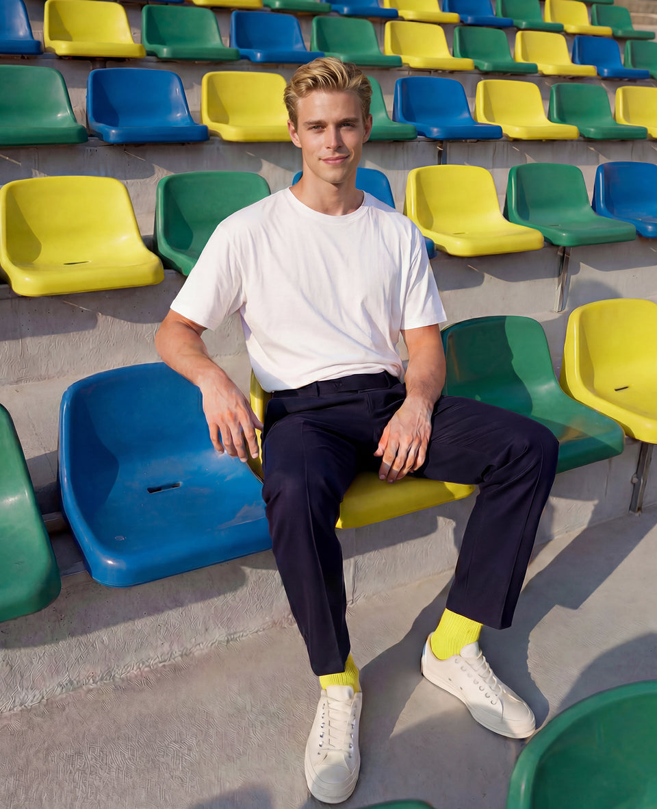 Man wearing Democratique Socks Originals Fine Rib in yellow with navy trousers and white sneakers in a football stadium with yellow, green, and blue seats — premium organic cotton supporter socks for Brazil and other national team colours.