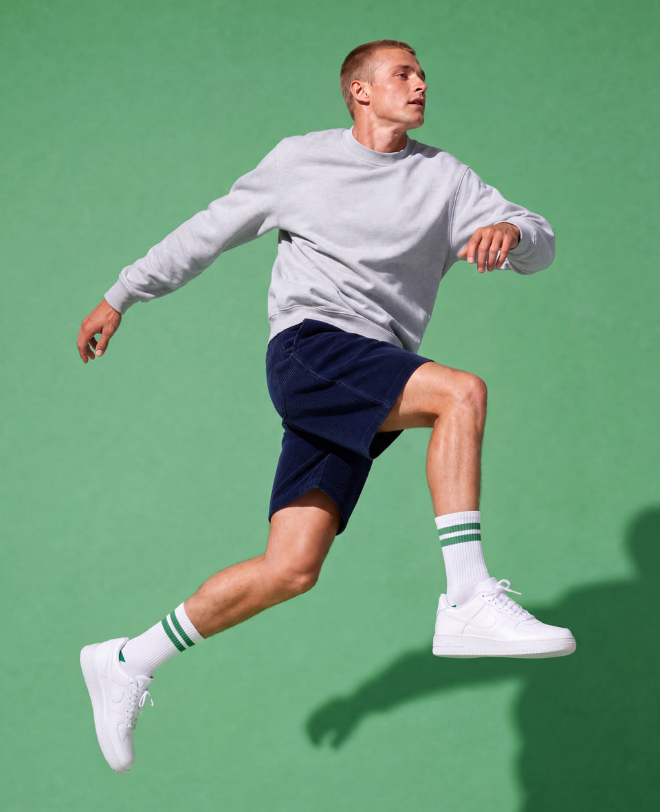 A man in a light gray sweatshirt, navy shorts, white socks, and white sneakers is captured mid-jump against a solid green studio background.