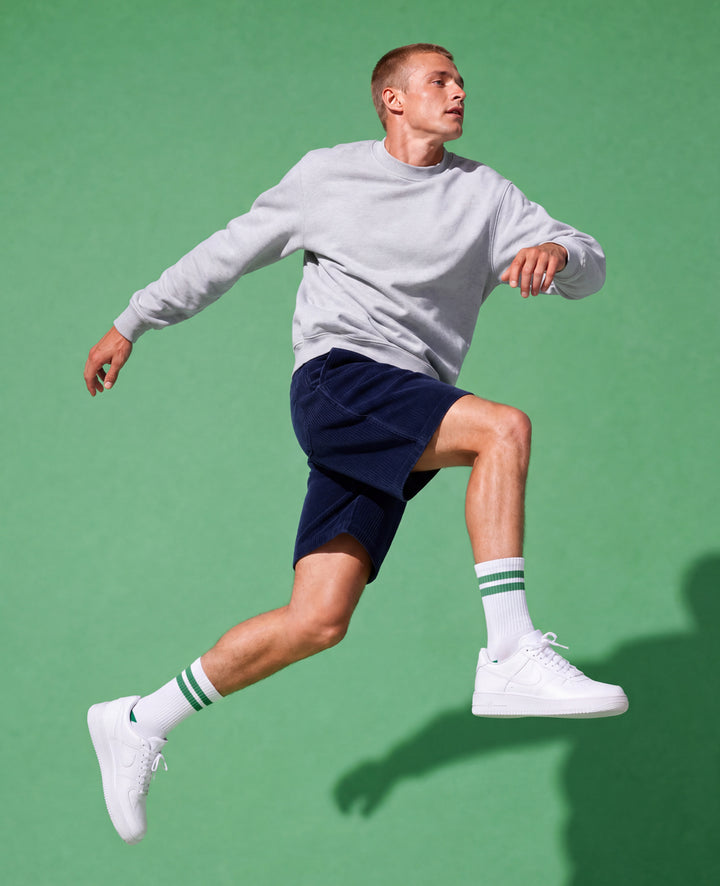 A man in a light gray sweatshirt, navy shorts, white socks, and white sneakers is captured mid-jump against a solid green studio background.
